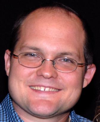 Erik Loebl smiling for the camera wearing a blue and black plaid collared shirt and glasses