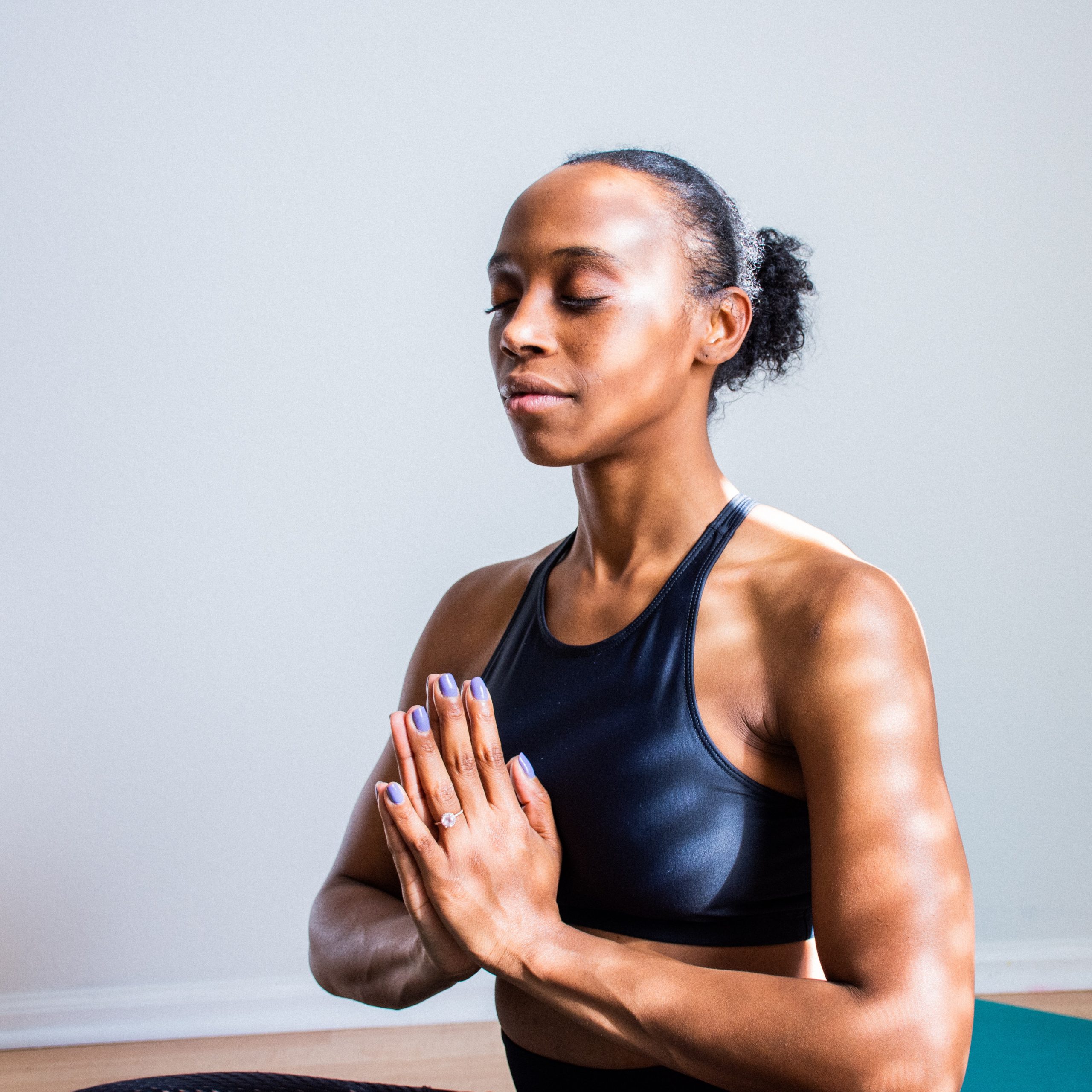 A melanated women with her hands in prayer and her eyes are closed in a yoga/meditation setting