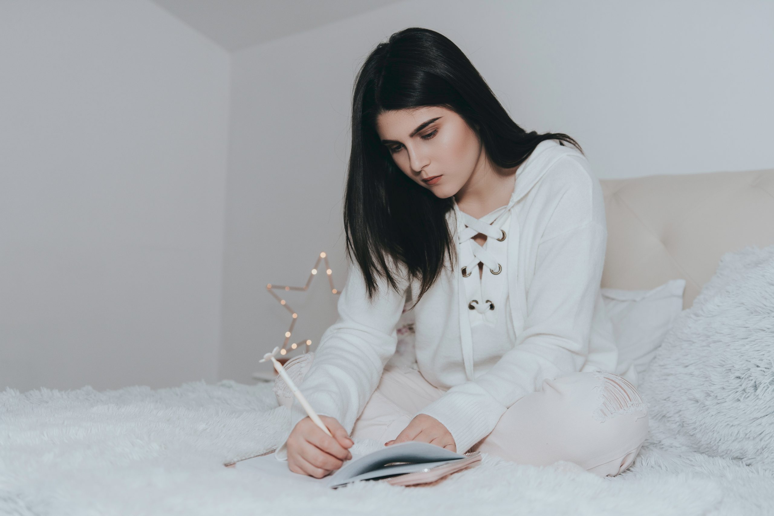 Woman with black hair wearing a white hoodie, on a white bed, in a white room, while writing in a journal