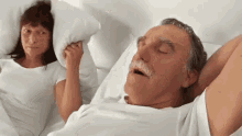 Husband snoring in white clothes and bedding, wife tries to smother him with a pillow