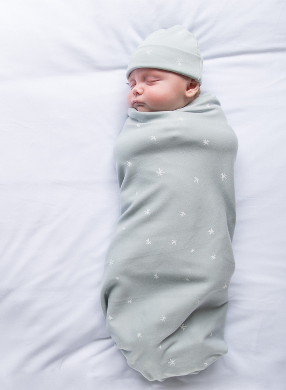 Small baby swaddled in a mint green blanket with matching beanie, lying on his back on a white bed.