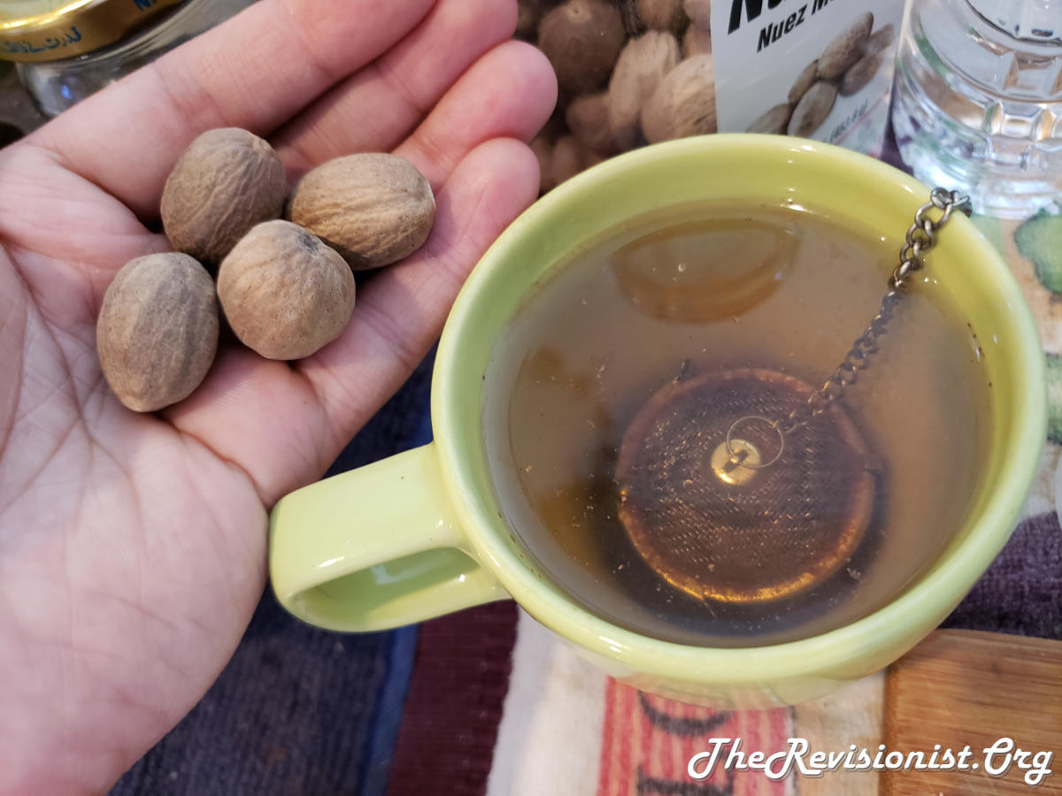 Nutmeg Tea in a yellow mug and someone is holding whole nutmeg seeds next to it