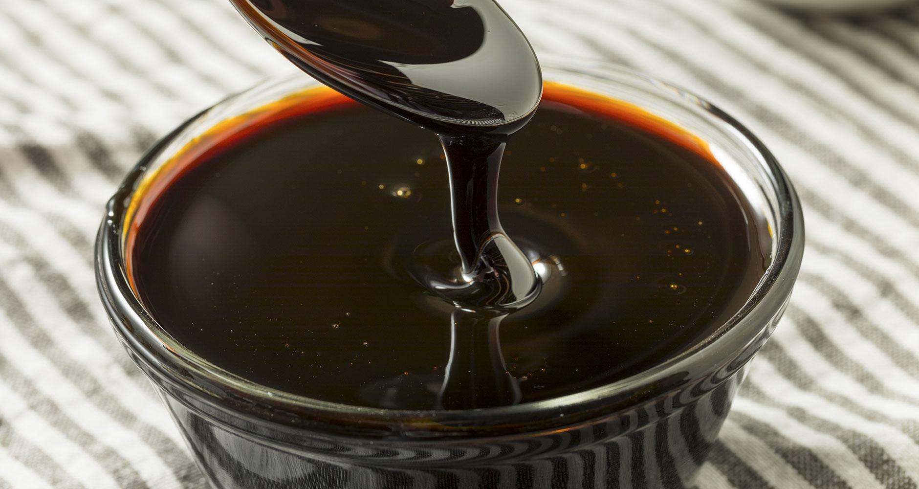 Blackstrap Molasses in a glass container getting scooped out by a silver spoon on a striped tablecloth