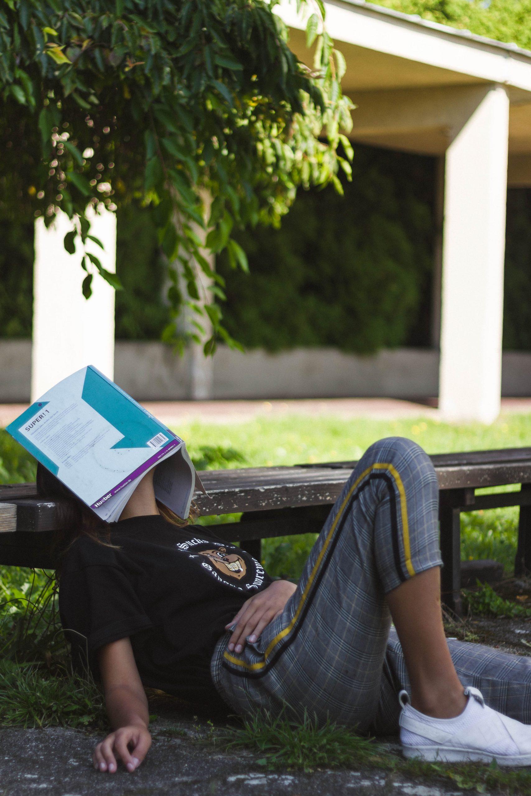 Woman on school campus leaning on a bench with a book covering her face, she is tired and the trees are green.