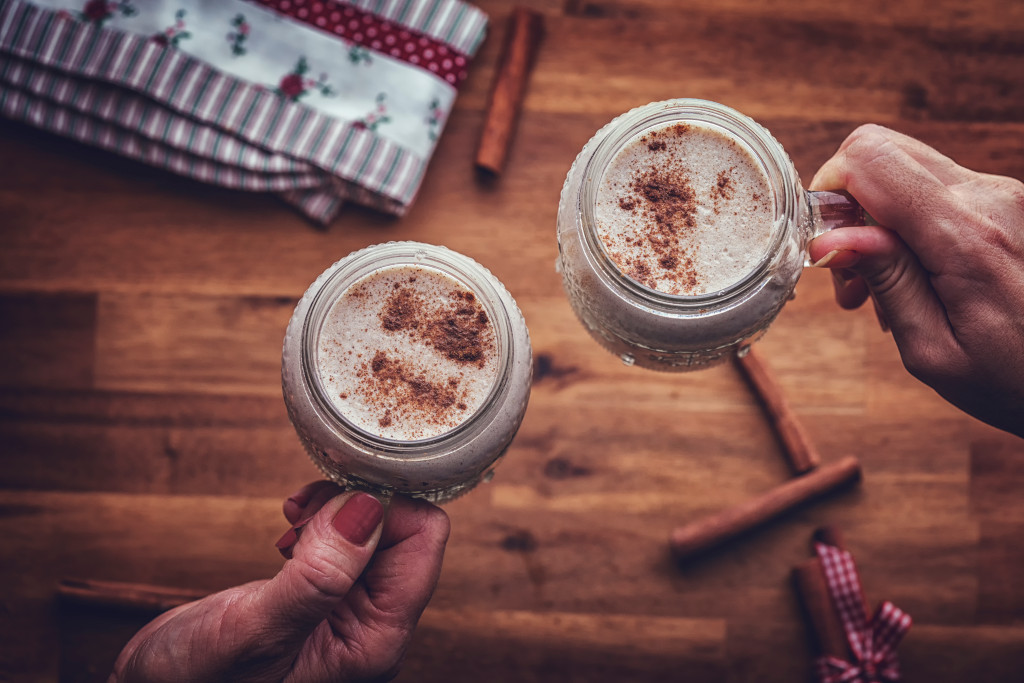 Two women's hands holding two different mugs with warm milk and nutmeg inside over a wooden table with cinnamon sticks on it